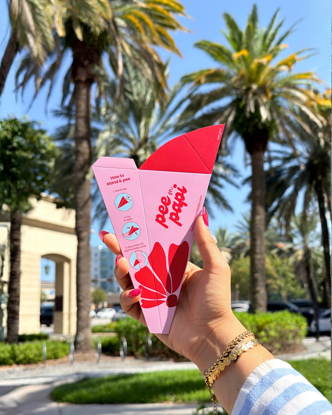 A hand holding a pink and red disposable pee funnel called Pee on Papi product package outdoors in front of palm trees and a sunny blue sky.