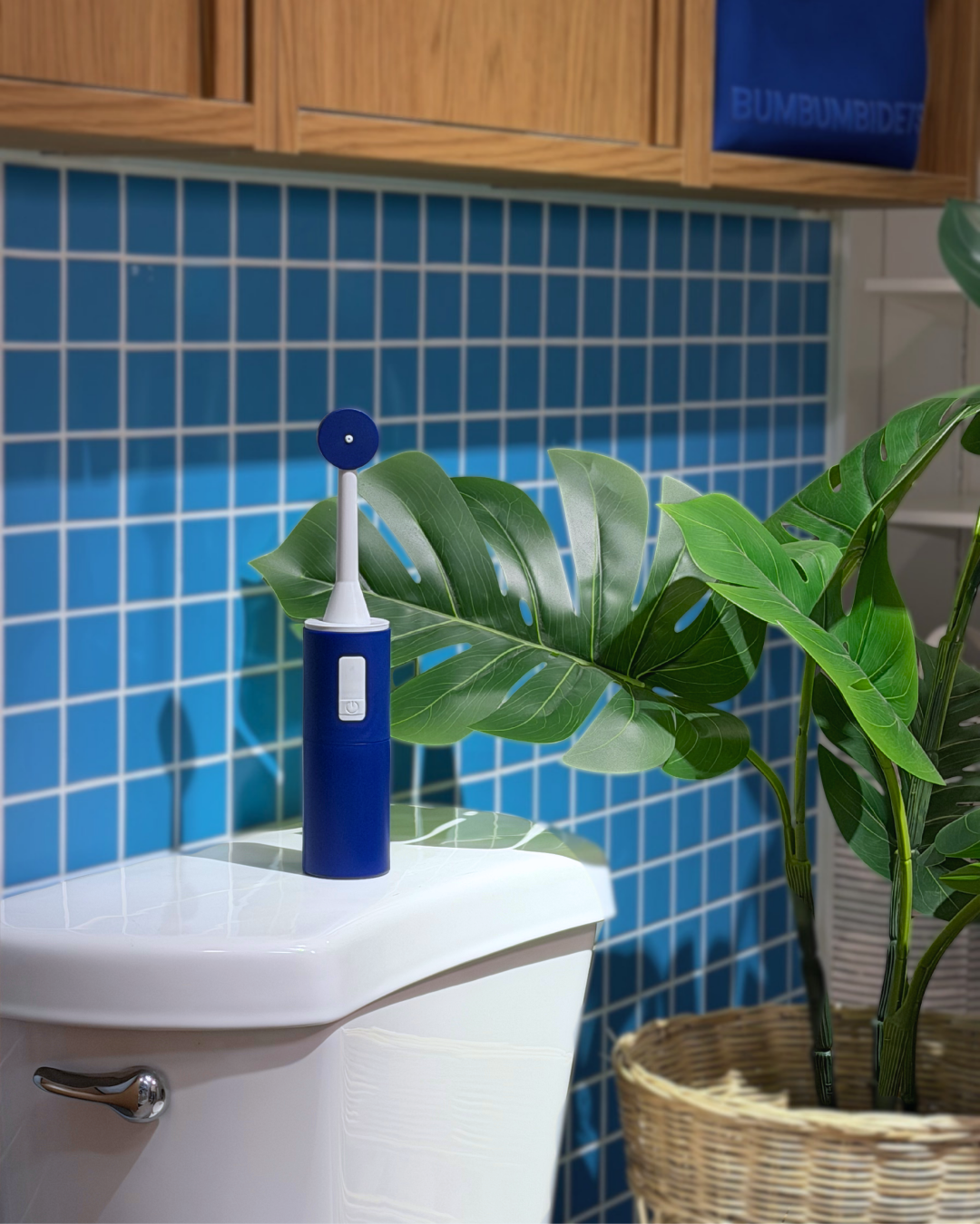 A blue and white electric portable bidet sits on the edge of a white toilet tank in a bathroom setting with blue mosaic tiles, wooden cabinets, and a green potted plant.