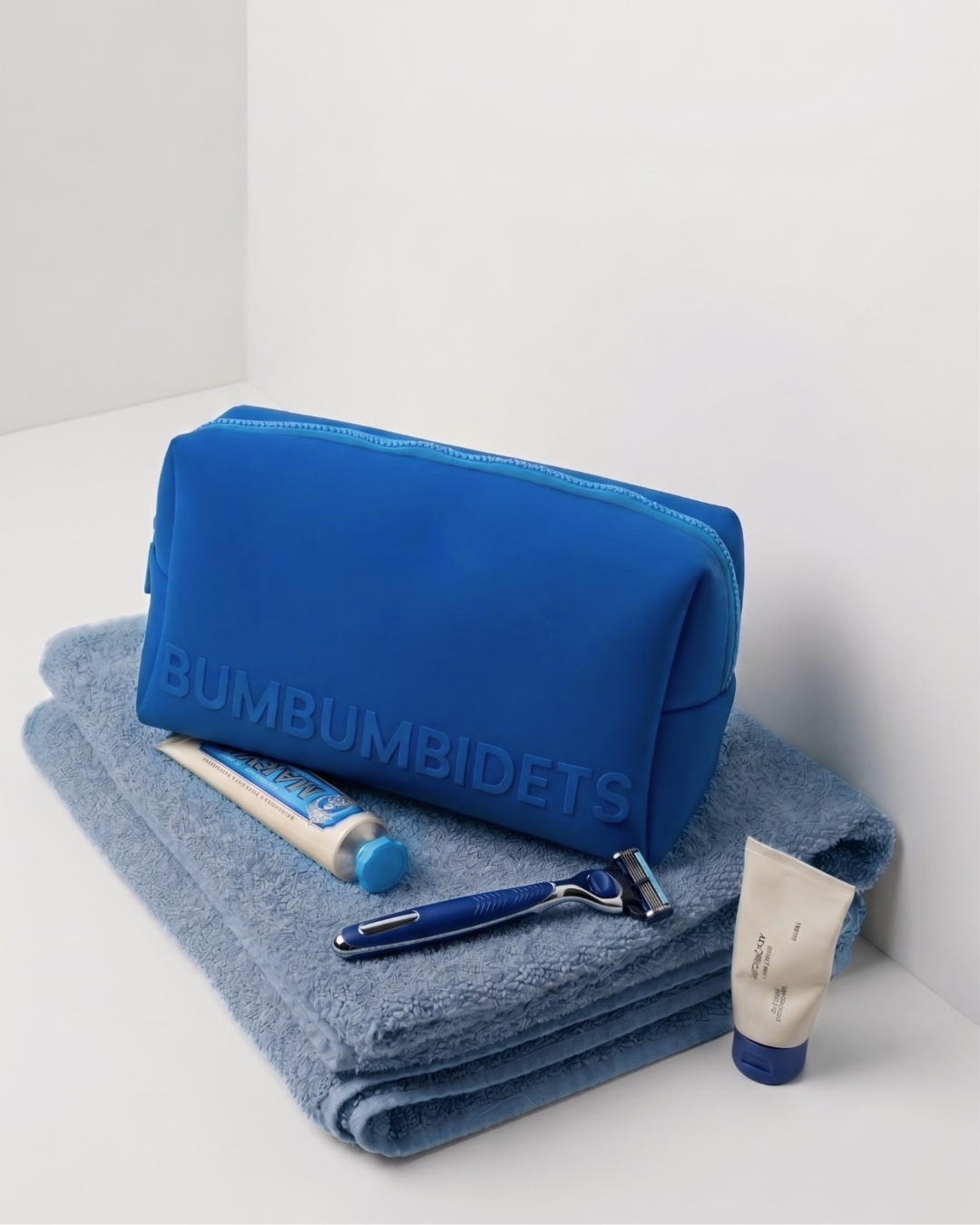 A blue toiletry bag with the brand name 'BUMBUMBIDETS' embossed on the front, displayed on top of a folded blue towel alongside a razor, a tube of toothpaste, and a small cream tube against a clean white background.