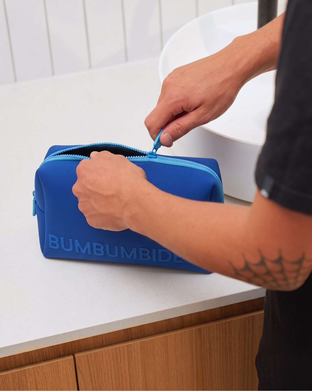 A person's hands unzip a bright blue neoprene zippered pouch with the brand name 'BUMBUMBIDIE' printed on the front, set on a white bathroom countertop.