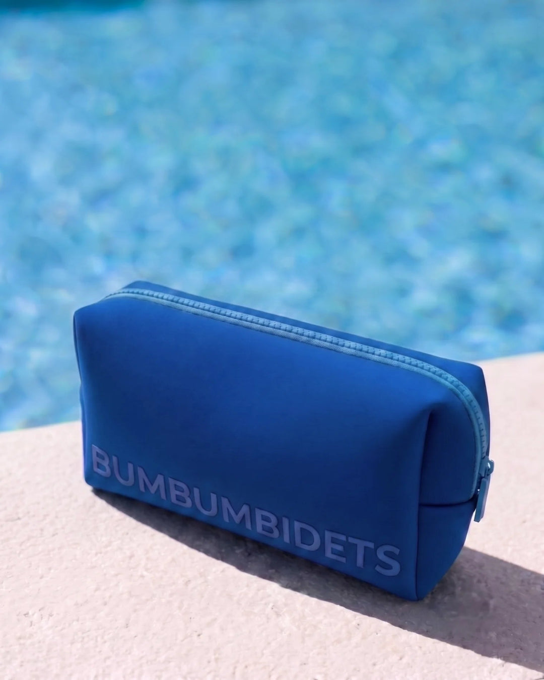 A blue zippered pouch with the brand name 'BUMBUMBIDETS' printed in light blue on the front, resting on a light-colored surface with a blurred swimming pool in the background.