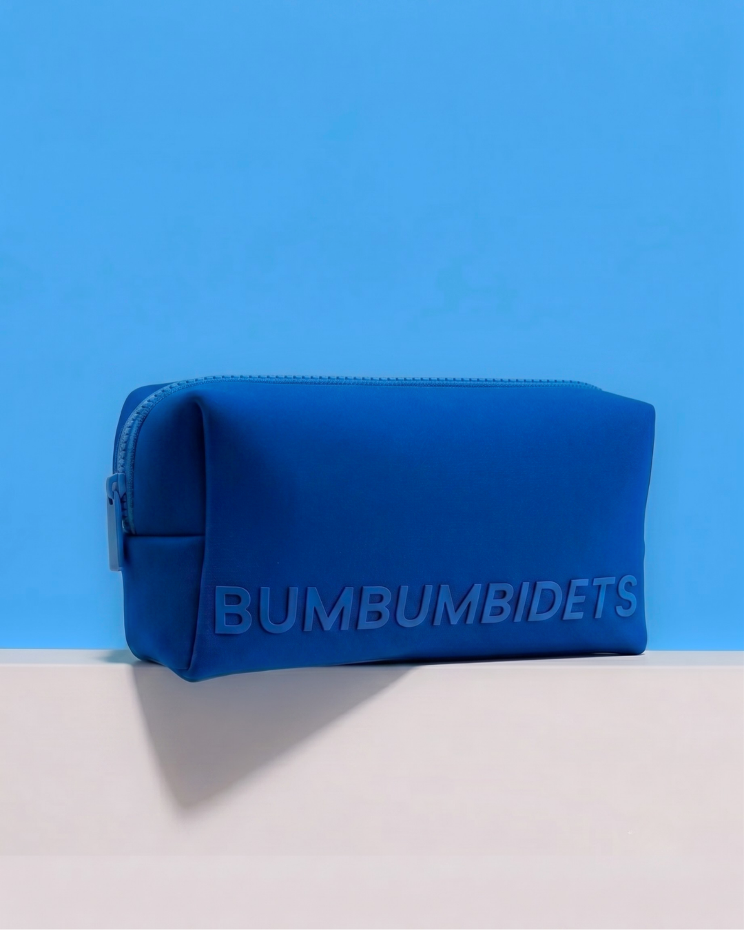 A blue zippered pouch with the brand name 'BUMBUMBIDETS' embossed on the front, resting on a white surface against a solid blue background.