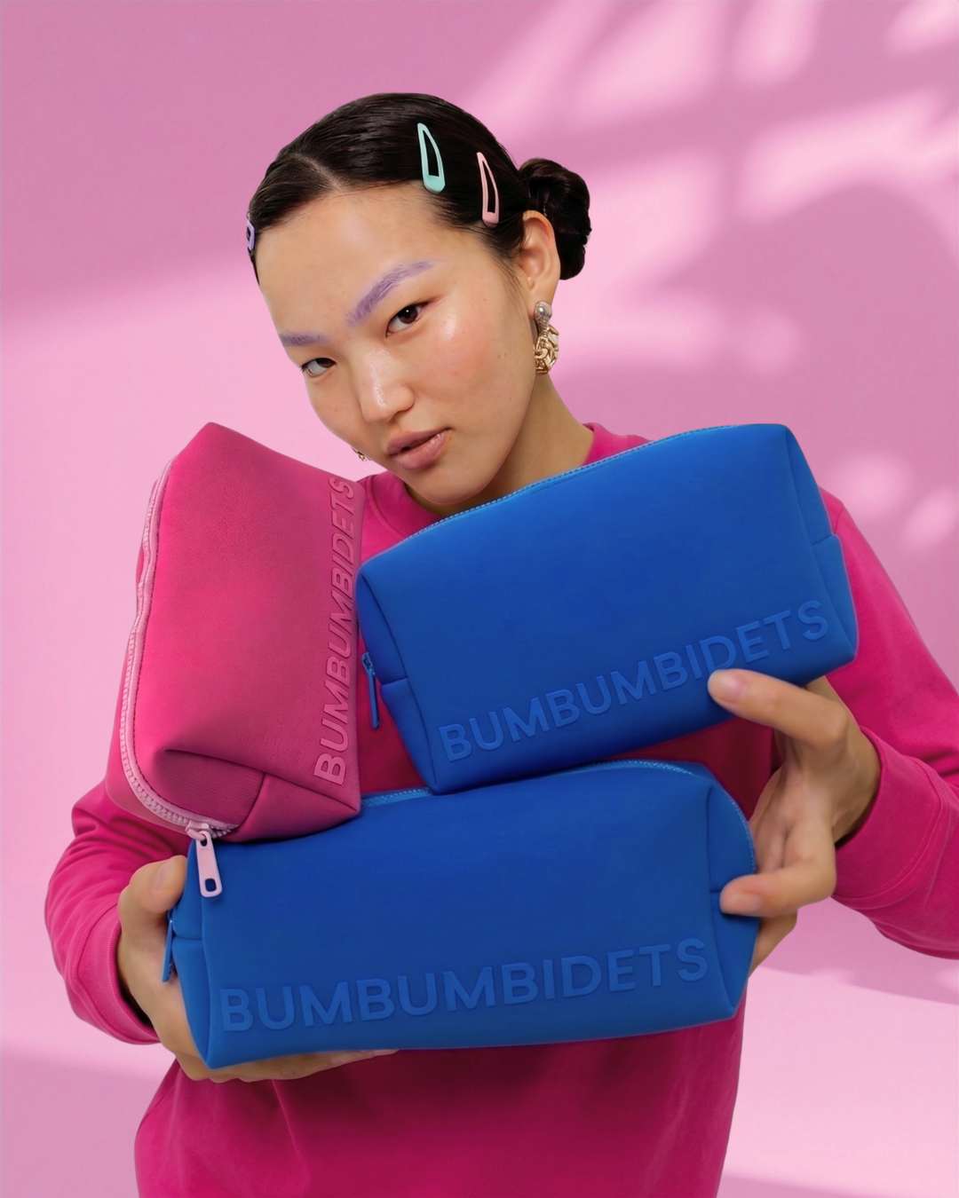 A girl wearing a pink sweatshirt holds three Bumbumbidets zippered pouches in pink and blue against a pink background with leaf shadows.
