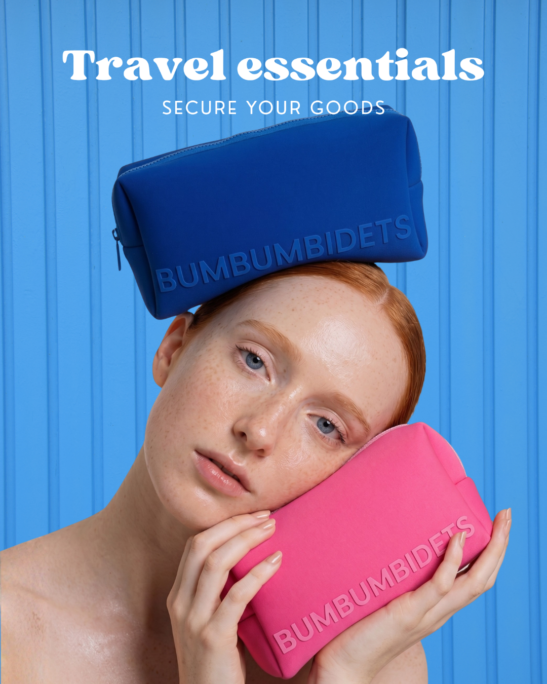 A model with red hair holds a pink BUMBUMBIDETS travel pouch against their face while wearing a blue version of the same pouch balanced on their head, set against a bright blue vertical-paneled background with the text 'Travel essentials SECURE YOUR GOODS'.