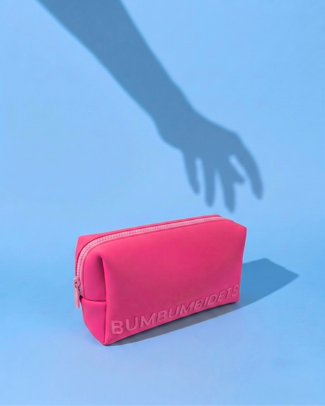 A bright pink zippered pouch with the brand name 'BUMBUMBEIDITS' embossed on the front, set against a light blue background with a hand shadow cast above it.