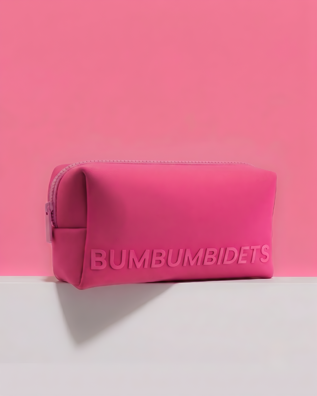 A bright pink zippered pouch with the brand name 'BUMBUMBIDETS' embossed on the front, set against a matching pink background and a white surface.