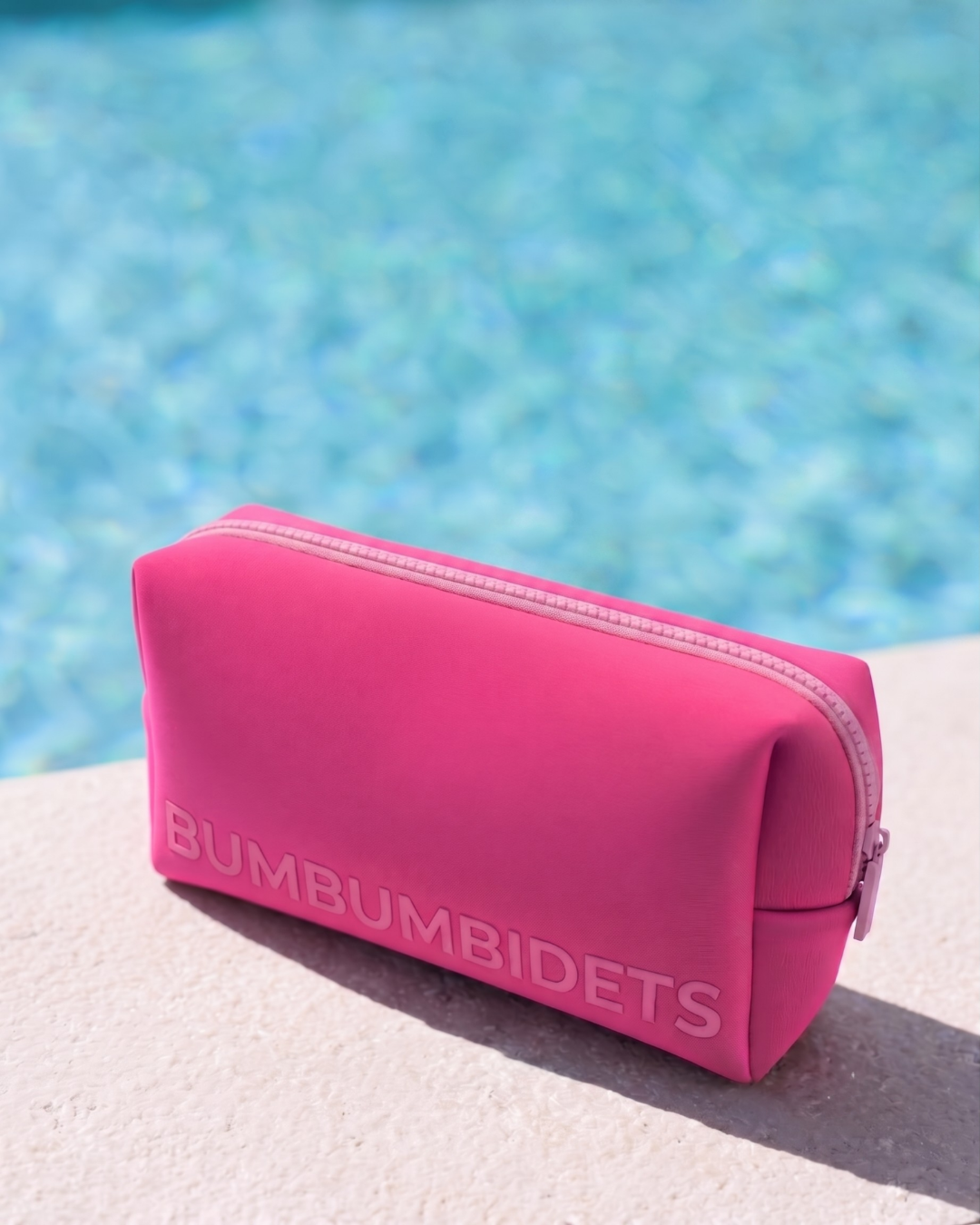 A bright pink zippered pouch with the brand name 'BUMBUMBIDETS' embossed on the front, resting on a light stone surface with a blurred swimming pool in the background.