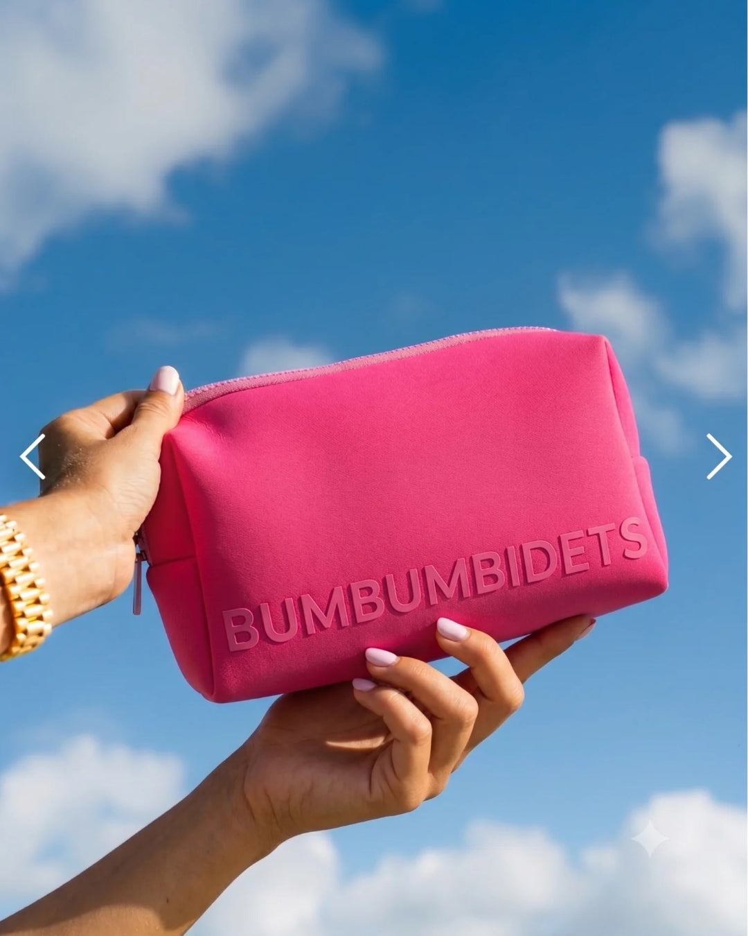 A bright pink cosmetic pouch with the brand name 'BUMBUMBIDETS' embossed on the front, held by two hands against a blue sky with clouds.