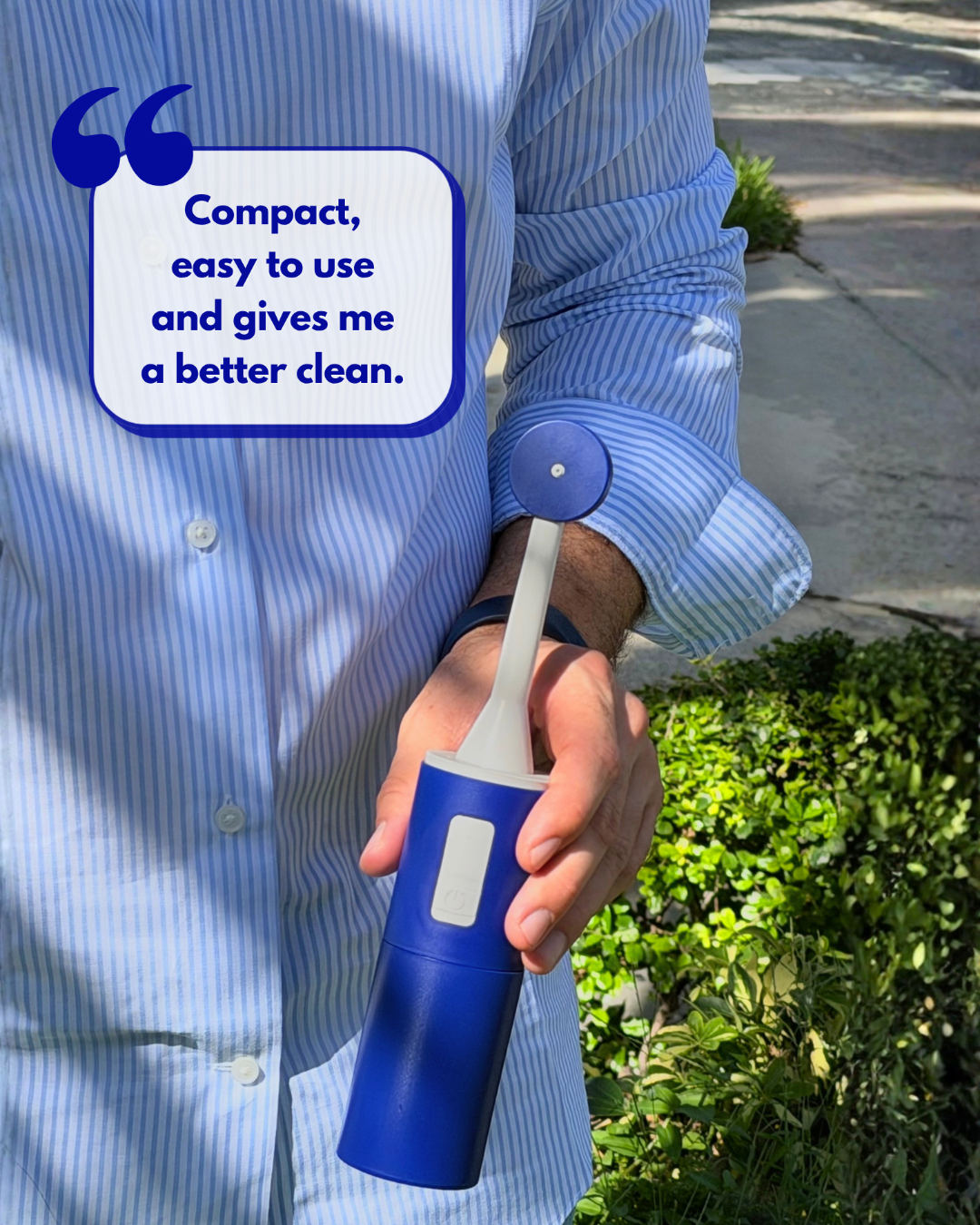 A person in a blue striped button-up shirt holds a compact blue handheld portable bidet outdoors near green shrubs and a sidewalk. A quote bubble reads, 'Compact, easy to use and gives me a better clean.